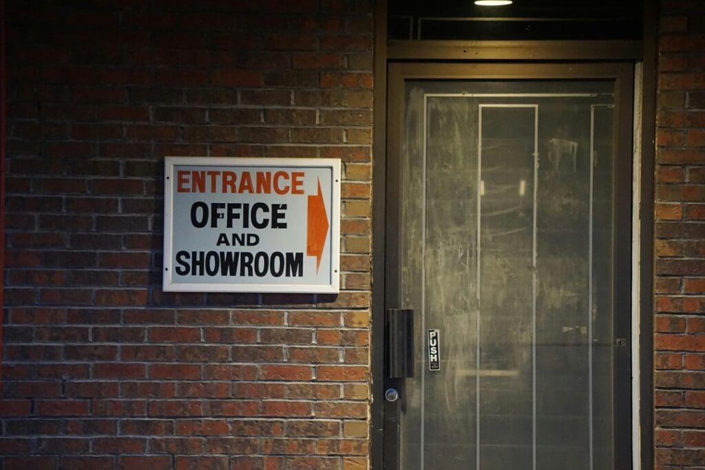 Office Signs Brick wall with a sign reading "Entrance Office and Showroom" with an arrow pointing right. Next to a glass door with a push handle.