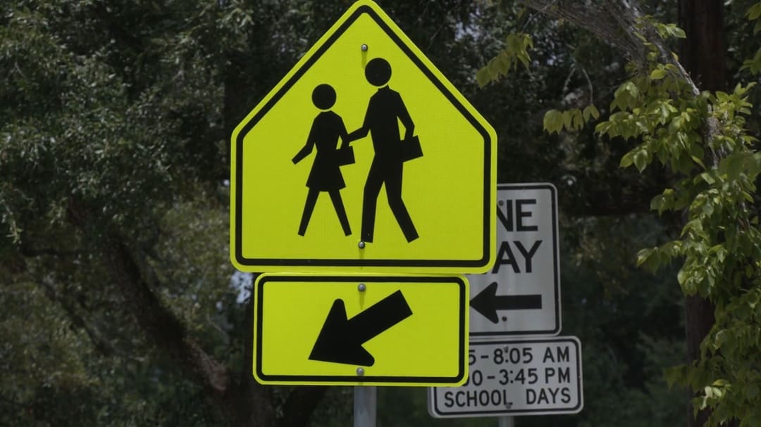 School Signs Nashville A yellow pedestrian crossing sign with silhouettes of two people walking. An arrow points left. A standard traffic sign with time restrictions is partially visible behind. Trees in the background.