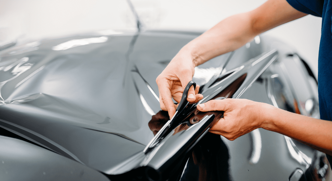 Person cutting a sheet of black vinyl wrap with scissors, applying it to the surface of a car.