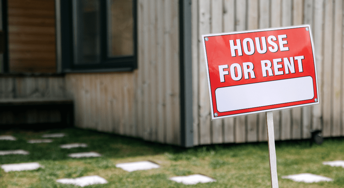 A red and white "House For Rent" sign is posted in front of a wooden house with a grass lawn and stepping stones.
