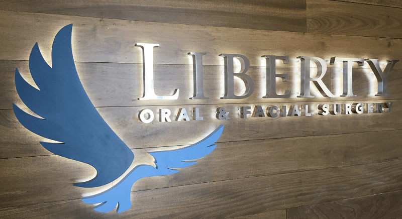 Wall sign for Liberty Oral & Facial Surgery featuring large metallic lettering and a blue eagle logo on a wooden background.