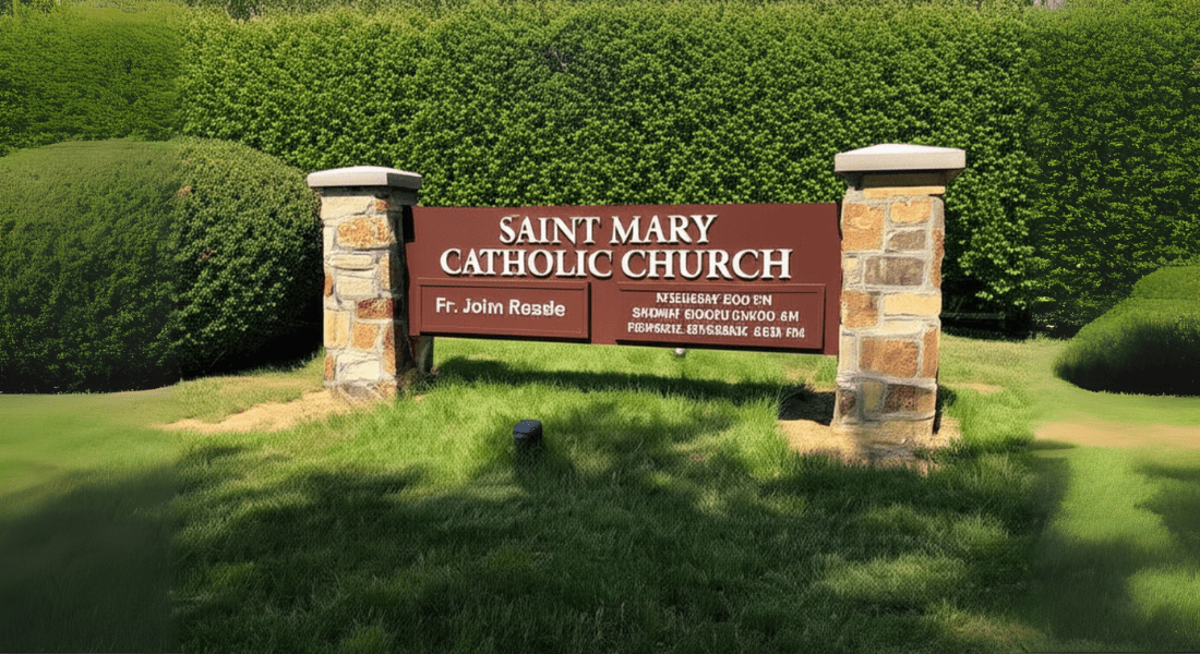 A brown sign with white text reads "Saint Mary Catholic Church Fr. John Rasle" and stands on a lawn between two stone pillars, with hedges in the background.