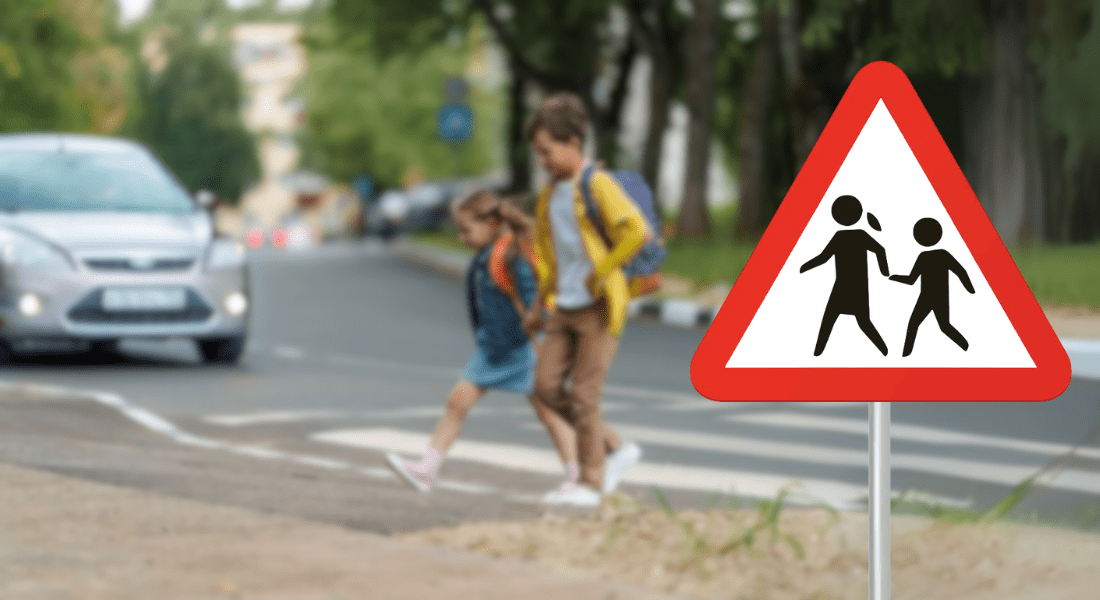Two children cross a street at a crosswalk near a triangular pedestrian crossing warning sign; a car approaches in the background.