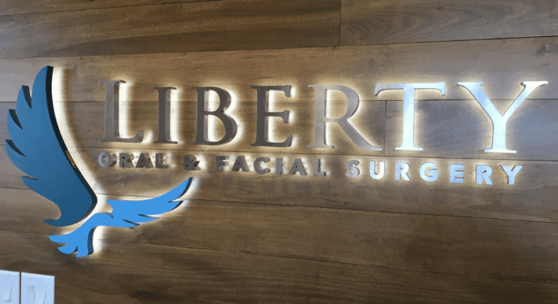 A wall sign reads "Liberty Oral & Facial Surgery" with a blue and white stylized wing logo on a wooden background.
