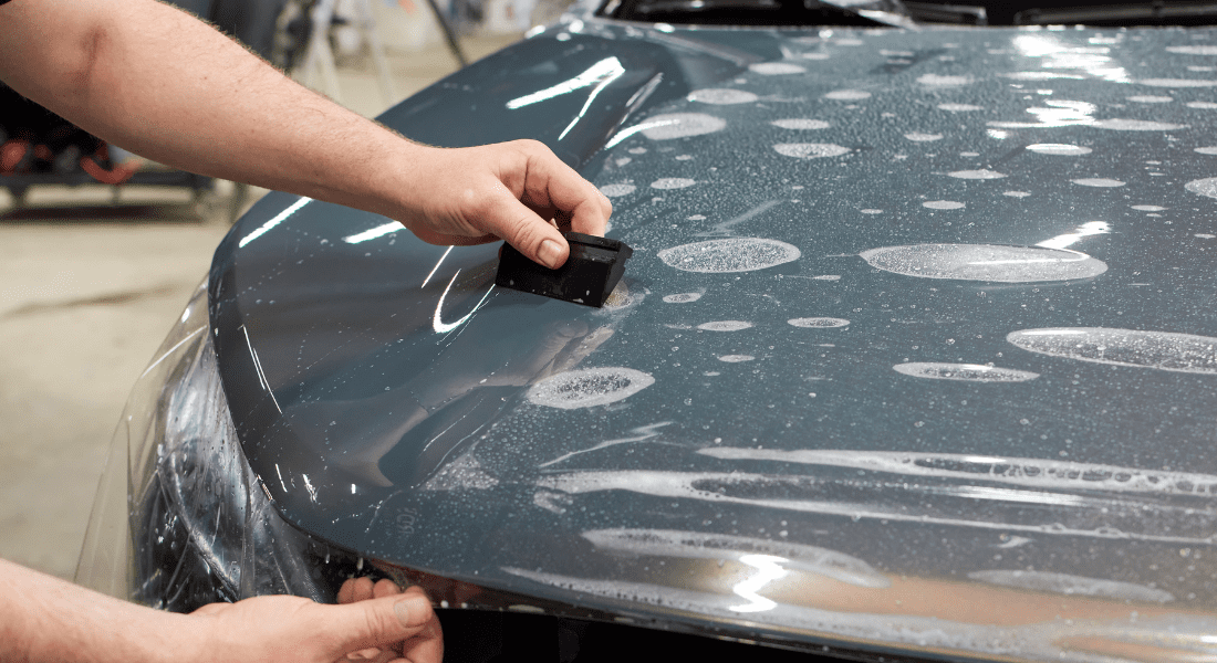 A person applies a protective film to a car hood, smoothing it with a tool while water spots are visible on the surface.