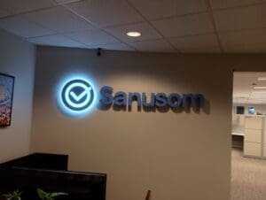 Illuminated "Sanusom" logo with a checkmark symbol on an office wall; cubicles and ceiling lights visible in the background.