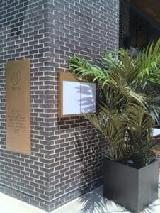 A bronze plaque and a blank sign are mounted on a brick building corner next to a large potted plant in sunlight.