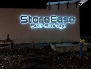 Illuminated "StoreEase Self-Storage" sign on an exterior wall at night, with exposed ground and utility pipes visible in front.