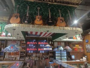 A bar interior with guitars hanging from the ceiling, an American flag, neon signs, and various bottles displayed behind the counter.