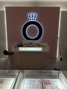 A jewelry display counter with assorted items in trays, a barstool, and a large illuminated circular logo on the wall behind. A broom is visible against the wall to the right.