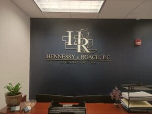 Reception area of Hennessy & Roach, P.C. law firm, featuring a wall sign with the firm&rsquo;s logo and name, a desk with a plant, mail trays, and office supplies.