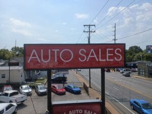 A large red "AUTO SALES INC." sign on a metal post is shown above a parking lot with several cars and a busy street in the background.