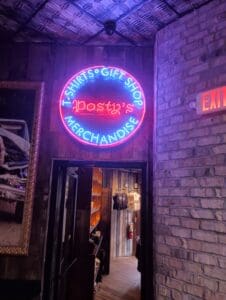 Neon sign reading "T-Shirts &bull; Gift Shop &bull; Merchandise Posty's" above a doorway leading to a room with racks of clothes. Brick wall and EXIT sign are visible.