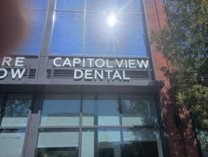 The front of a building with large windows and a sign that reads "CAPITOL VIEW DENTAL" in white letters; sunlight reflects off the glass.