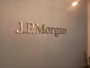Metallic "J.P. Morgan" letters are mounted on a plain light gray wall, with part of a light fixture and a plant visible at the edges.