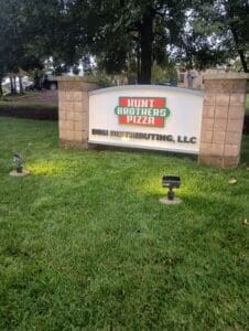A white and tan outdoor sign reads "Hunt Brothers Pizza" and "DBH Distributing, LLC," positioned on a grassy area with two small spotlights.