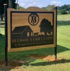 A wooden sign reads &ldquo;Bledsoe Family Farm, A Tennessee Century Farm&rdquo; with a barn, tree, cow, and the text &ldquo;Est. 1898&rdquo; engraved above. The sign stands on grassy land.
