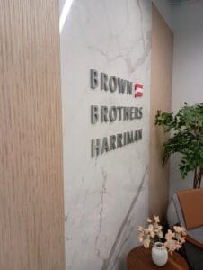 Brown Brothers Harriman sign on a marble wall, next to a plant and a small table with a flower vase in an office setting.