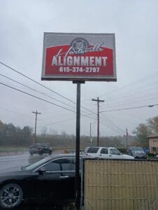A red and white sign for "Hartsville Alignment" with a phone number above a street and parked cars on a rainy day.
