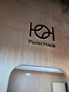 A wood-paneled wall with a black illuminated "Padel Haus" logo and text above an arched doorway.