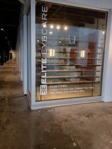 Storefront window for Elite Eyecare with glasses displayed on shelves inside; exam types and services are listed on the window.