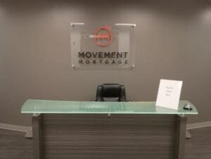 Reception desk with a glass top, a black chair behind it, a sign reading "PLEASE RING BELL," and a bell. "MOVEMENT MORTGAGE" logo is displayed on the wall.