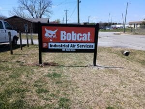 A sign for Bobcat Industrial Air Services at 138 Balm Drive stands on a grassy area near a road and a white truck.
