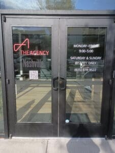 Glass doors of "The Agency Nashville" with business hours, contact number, QR code, and "By Appt Only" note for weekends displayed in white text.