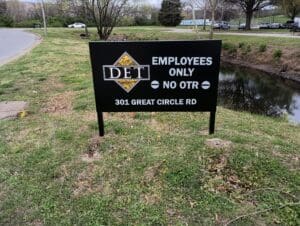 A black sign reads "DET Employees Only = No OTR, 301 Great Circle Rd" and stands on grass near a road and a pond.
