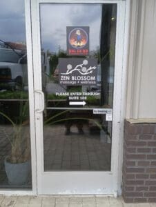 Glass door with signs for "Big Burd Fitness and Wellness" and "Zen Blossom Massage + Wellness," and a notice to enter through Suite 105; "No Smoking" sign and reflection visible.