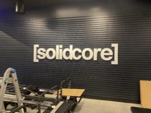 A wall with the word &ldquo;[solidcore]&rdquo; in large white letters; fitness equipment and a ladder are visible in the foreground.