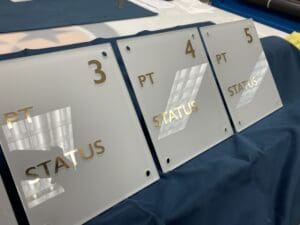 Three white acrylic plaques labeled "PT STATUS" with gold numbers 3, 4, and 5 rest on a dark blue surface. Each plaque has four mounting holes in the corners.