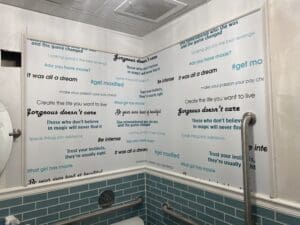 White bathroom walls feature motivational phrases in black and blue text. The restroom includes a metal grab bar, teal tile, and part of a round mirror is visible on the left.