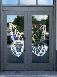 A set of glass double doors, each featuring half of a white butterfly design that forms a complete butterfly when the doors are closed. Buildings and trees are visible outside.