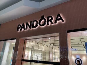 Storefront of a Pandora jewelry shop with illuminated black lettering on a pink tiled wall and a partially closed metal security gate.