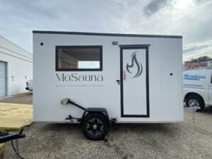 A white mobile sauna trailer labeled "MoSauna" with a window, door, and fire flame logo, parked on concrete near a building and a white van.
