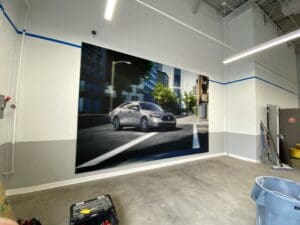 A large wall mural of a silver car driving in a city is displayed inside an industrial-style room with tools and cleaning supplies visible.