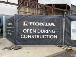 A large black banner at a Honda dealership reads "Honda Open During Construction" in white letters, with construction materials and fencing visible.