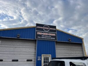 A blue automotive repair shop building with a sign showing the business name, phone number, address, and "Full Service Auto Repair." A white vehicle is parked in front.