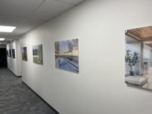 Framed photographs of modern architecture and outdoor scenes are displayed on a white hallway wall with gray carpeting and a drop ceiling.