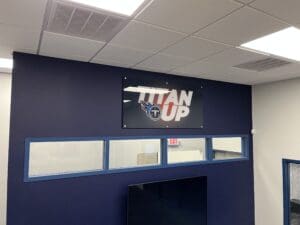 A sign reading "TITAN UP" with the Tennessee Titans logo is mounted on a dark blue wall above windows in an office space.