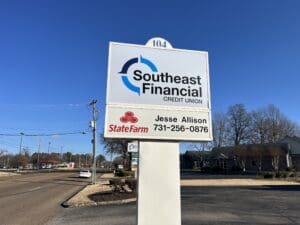 A roadside sign displays Southeast Financial Credit Union and State Farm with agent Jesse Allison&rsquo;s contact number, against a clear blue sky and nearby buildings.