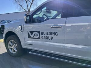 White pickup truck parked in sunlight with "VP Building Group" logo and text on the driver's side door.