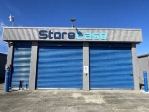 A storage facility with three blue roller doors, gray walls, and "StoreEase" sign above. The area is empty and lit by sunlight.