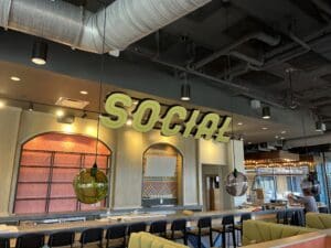 A modern bar interior with a large "SOCIAL" sign hanging from the ceiling, rows of bar stools, and decorative lighting fixtures.