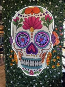 Large decorative sugar skull with colorful floral patterns is mounted on a green leafy wall, featuring vibrant flowers filling the eyes, nose, and cheeks.