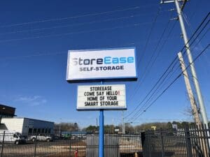 A StoreEase Self-Storage sign with a marquee that reads, "STOREEASE COME SAY HELLO! HOME OF YOUR SMART STORAGE" stands near a fenced lot under a clear blue sky.