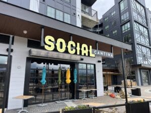 Exterior of Social Cantina restaurant with closed umbrellas, stacked chairs, and outdoor tables in front of a modern apartment building.