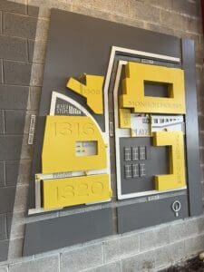 A mounted 3D map with yellow and gray blocks displays labeled buildings: River Steps, Core, Monroe House, Passage, and Plaza, on a cinder block wall.