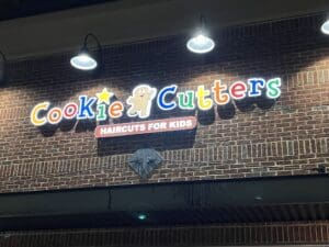 A brightly lit sign on a brick building reads "Cookie Cutters" with a gingerbread figure and the tagline "Haircuts for Kids" below.